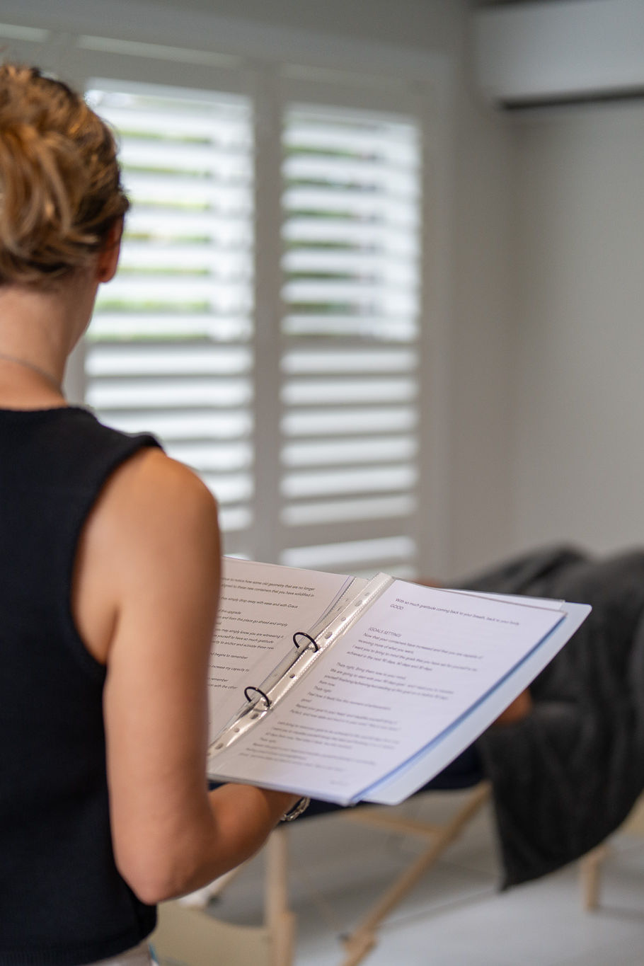The therapist is holding a clipboard with a hypnotherapy program on it, in the background is a patient lying on their back on a massage table.